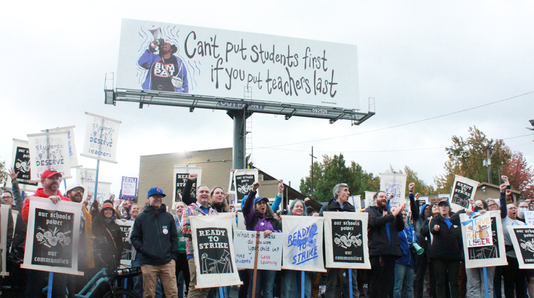Portland Public School workers and community members with signs in support of the teacher's union stand below a billboard that says, "Can't put students first if you put teachers last."