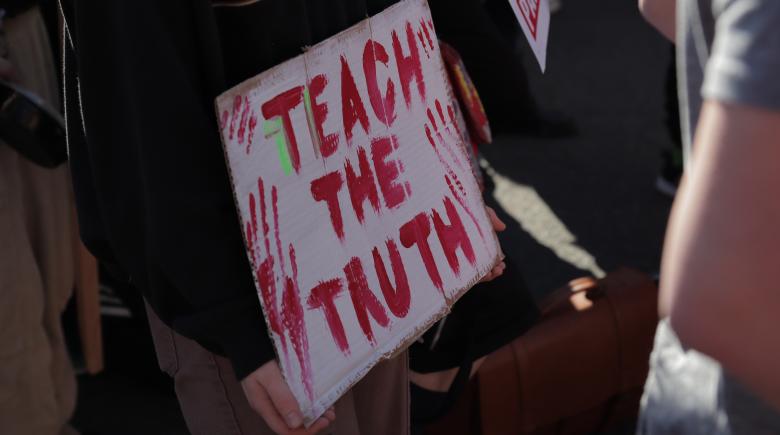 A pair of hands hold a cardboard sign that says, "teach the truth."