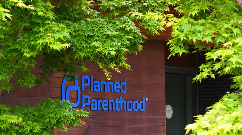 A blue Planned Parenthood sign on the side of a building is surrounded by green leaves on a tree.