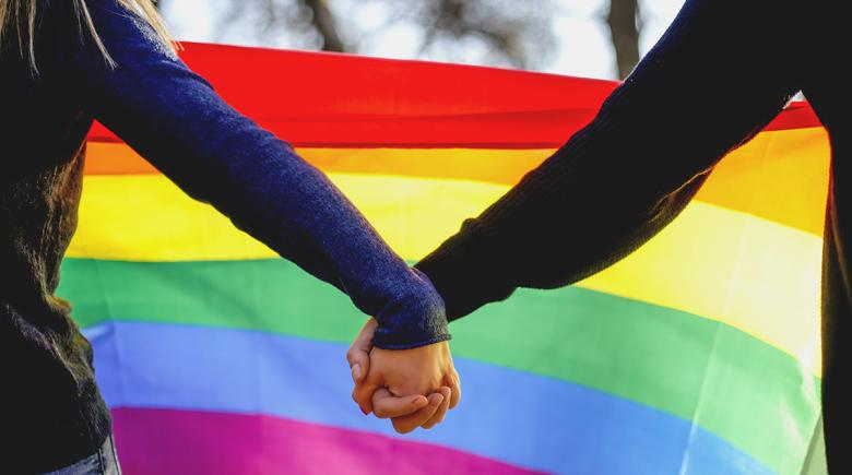 Two people hold hands in front of a rainbow Pride flag