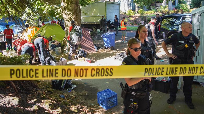 Tape that says "police line do not cross" sections off an area. In that area, three police officers are standing watch. People behind them are gathering possessions and tents on the sidewalk.