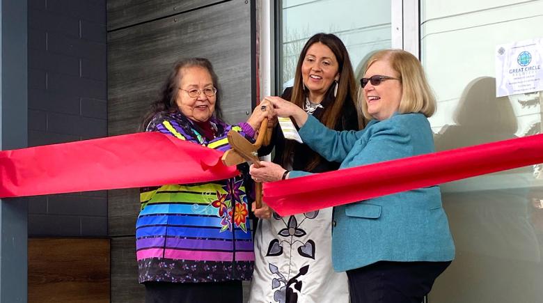 3 women are smiling and holding a large pair of scissors to cut a ribbon at a ribbon cutting ceremony for a new opioid treatment clinic in Portland. 