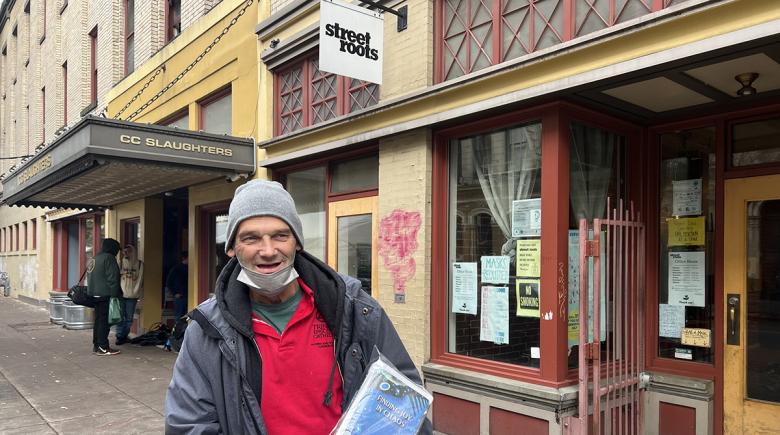 Rick Davis smiles for a photo and holds issues of Street Roots Holiday Zine while standing in front of the Street Roots offices.