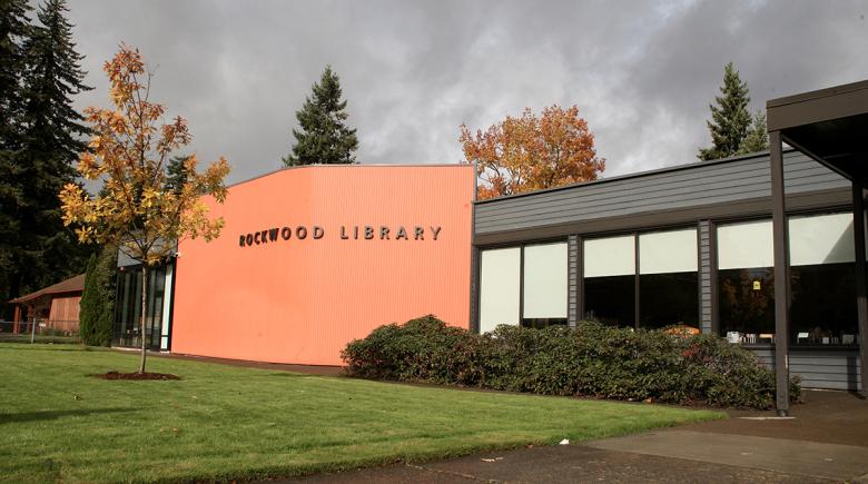 The exterior of the Rockwood Library. The building is partly red and gray with large windows. In front of the building is a green lawn with a tree.