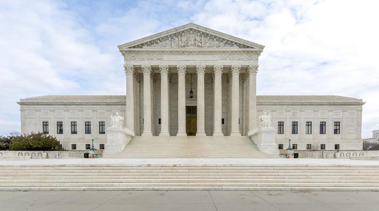 A blue sky in the background and in the foreground the Supreme Court of the United States building fills the photo. The building is white with tall pillars and stairs.