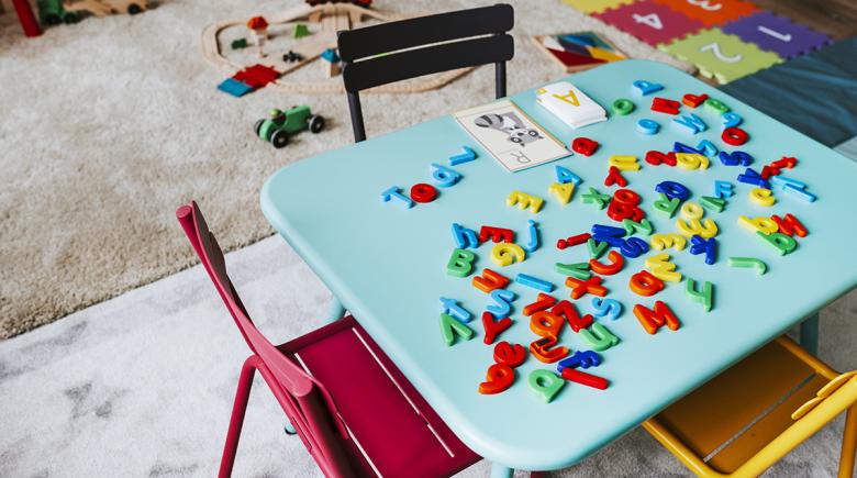 A stock photo of a preschool classroom. A table is filled with alphabet magnets and a rug with toys is in the background.