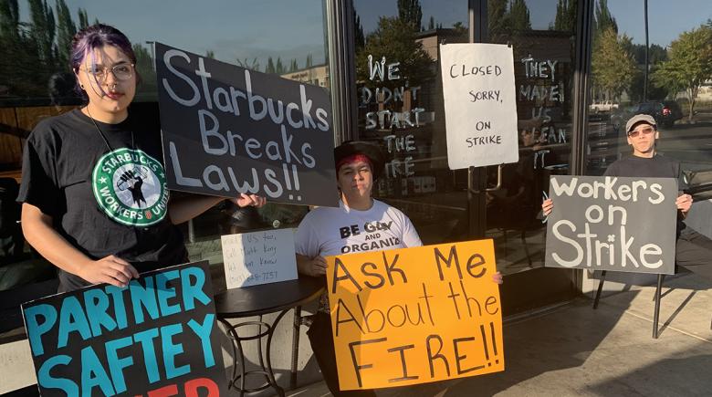 Three people hold signs outside of Starbucks. The signs say, "Starbucks Breaks laws. Closed, sorry on strike. Workers on strike."