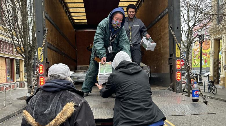 Four people are working to unload newspapers from the delivery truck. One person is handing off a bundle of newspapers to someone else outside the truck.