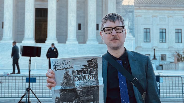 Jeremiah Hayden stands outside the Supreme Court holding up an issue of Street Roots.