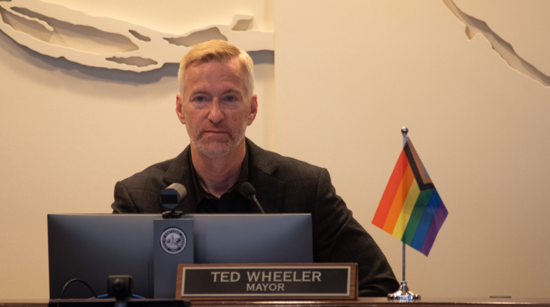 Ted Wheeler sitting in the city council chamber. He has a nameplate in front of him and a rainbow flag next to his nameplate.