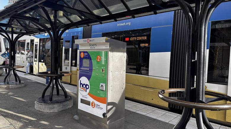 A TriMet MAX stop with a fare machine. In the background a MAX train approaches the stop.