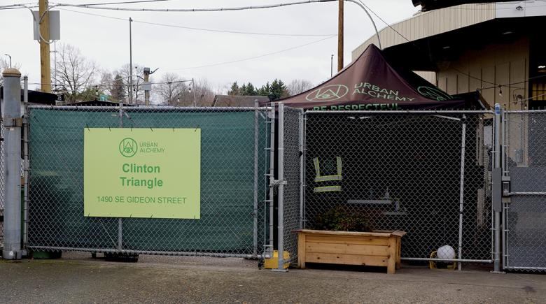 The outside of the Clinton Triangle site. It shows a fenced off area with a green sign that says "Urban Alchemy. Clinton Triangle."
