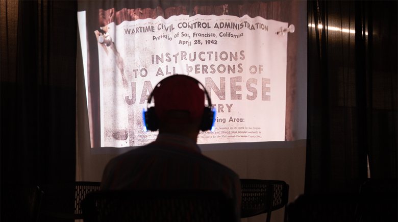 A person wears headphones and sits in front of a screen in a dark room. Large text on the screen says "instructions to persons of Japanese ancestry"
