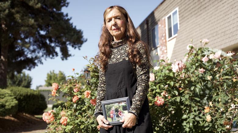 Vickie Hanson among rose bushes and she is holding a framed family photo in her hands. Vickie has long brown hair that sits on her shoulders.