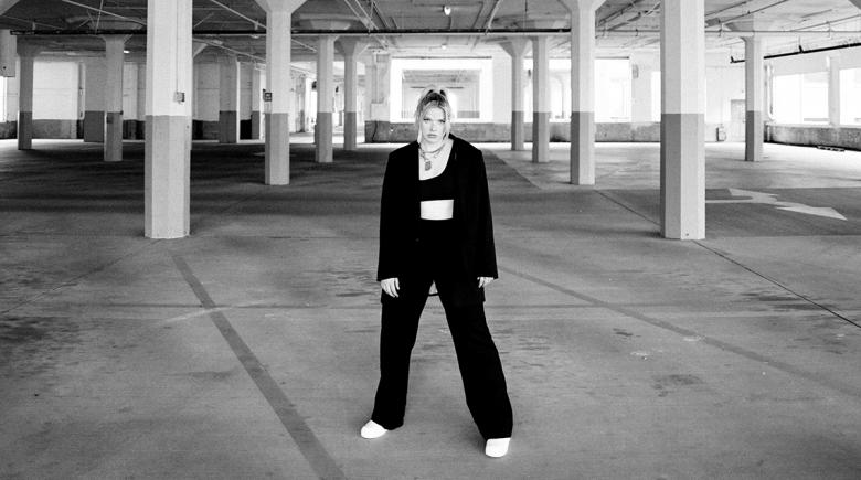 Photo of rapper Wynne surrounded by cement pillars in an empty building. She's wearing all black in a blazer and black pants and white shoes.