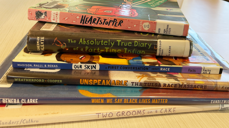 A stack of books sits on a table. Some titles include "Heartstopper, Two Grooms on a Cake."