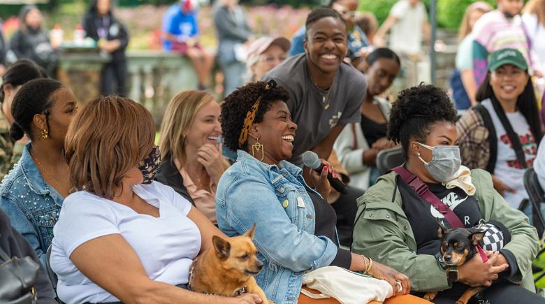 People sitting in the audience look to the stage as one person asks a question into a mic. Some attendees have their dogs in their lap. The people in the photo are smiling and laughing.