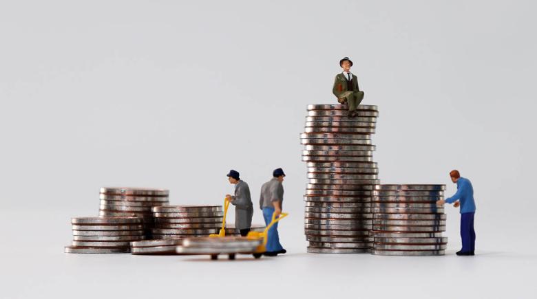 Miniature figurines depict workers moving coins around while one figurine man is in nicer clothes sitting atop a large pile of coins. The image evokes the contrast between high earners and low wage workers.