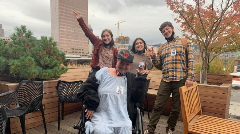 4 people smile and pose for a photo on a rooftop. The people are surrounded by chairs and in the background are trees and tall buildings in downtown Portland.
