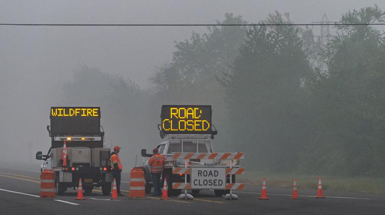 A highway is closed off with two signs. One sign says, “wildfire” and the other says “road closed.” The air is thick with smoke and two workers stand next to the trucks blocking off the road.